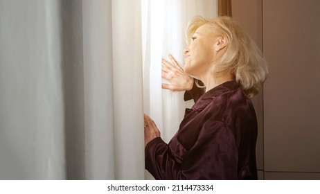 Mature Lazy Woman With Long Blonde Hairstyle And Bangs Stretches Arms After Waking Up And Looks Out Of Window Through Curtain Standing In Kitchen, Sunlight.