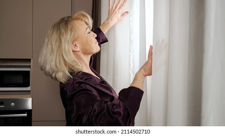 Mature Lazy Woman With Long Blonde Hairstyle And Bangs Stretches Arms After Waking Up And Looks Out Of Window Through Curtain Standing In Kitchen, Closeup.