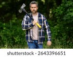 Mature Lumberjack with serious face holds axe in the forest. A lumberjack with an ax examines the tree before felling. Serious lumberjack. Millennial middle aged man holding a big axe outdoor.