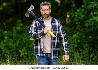 Mature Lumberjack with serious face holds axe in the forest. A lumberjack with an ax examines the tree before felling. Serious lumberjack. Millennial middle aged man holding a big axe outdoor.