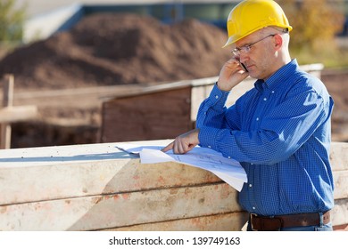 Mature Male Architect Pointing At Blueprint While Using Phone At Construction Site