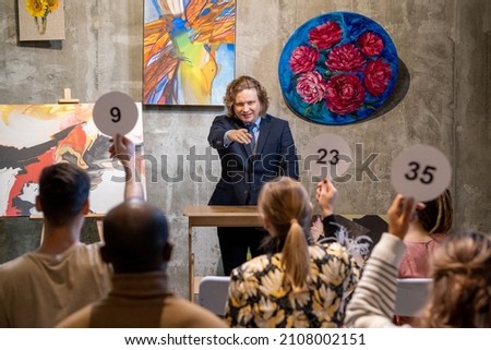 Mature male auctioneer in formalwear standing by tribune and pointing at one of people with auction paddles
