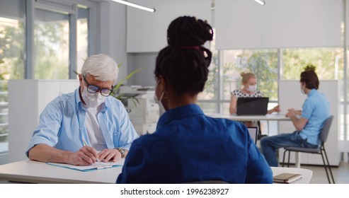 Mature Male Client In Medical Mask Sign Loan Agreement In Bank. Back View Of Afro-american Female Manager Consulting Aged Customer Wearing Protective Mask In Modern Office