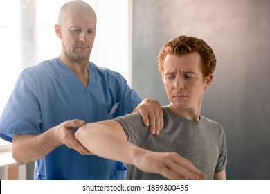 Mature Male Clinician In Blue Uniform Supporting Bent Arm Of Young Patient During Physiotherapeutic Training In Modern Rehabilitation Clinics