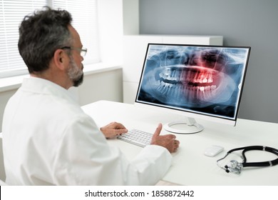 Mature Male Dentist Looking At Teeth X-ray On Computer In Clinic