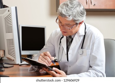 Mature Male Doctor Reading While Sitting In Front Of Computer At Desk In Clinic