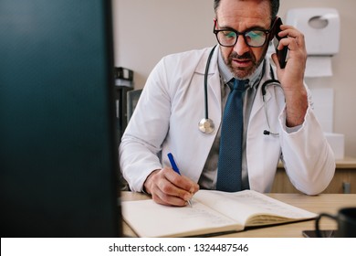 Mature Male Doctor Talking On Cell Phone And Making Notes In His Book. Physician Making Phone Call And Writing In His Diary.