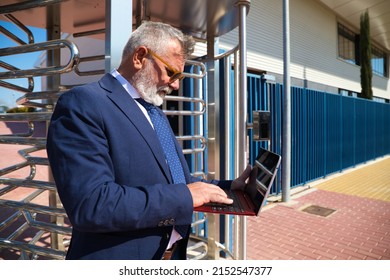 Mature Male Executive, Grey-haired, Bearded, With Sunglasses, Jacket And Tie Is Consulting His Laptop Computer In The Street. Concept Businessman And Executive.