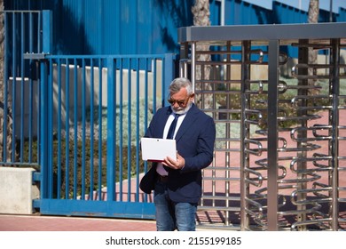 Mature Male Executive, Grey-haired, Bearded, With Sunglasses, Jacket And Tie Is Consulting His Laptop Computer In The Street. Concept Businessman And Executive.