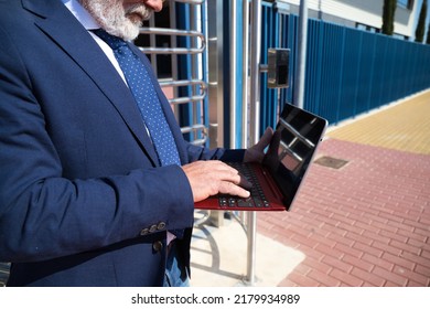 Mature Male Executive, Grey-haired, Bearded, With Sunglasses, Jacket And Tie Is Consulting His Laptop Computer In The Street. Concept Businessman And Executive.