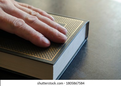 Mature Male Hand Rests On Top Of A Closed Book. Green Hardcover Book With Gold Lettering. Male Hand With Calluses And Cuts. Selective Focus.