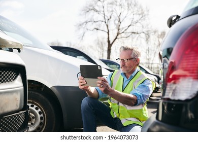 Mature Male Loss Adjuster Wearing Hi-vis Safety Vest Standing In Compound For Damaged Cars Taking Photo For Insurance Claim Report On Digital Tablet