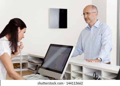 Mature male patient looking at female receptionist using landline phone and computer at reception in dentist's clinic