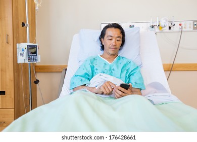 Mature Male Patient Text Messaging Through Cell Phone On Bed In Hospital