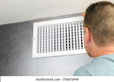 Mature male peering inside an upper wall white grid air duct on a silver wall near a white ceiling. A guy inspecting a heating and cooling air register duct for maintenance.
