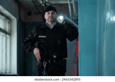 Mature male security guard in uniform and cap lighting forward with flashlight while moving along corridor of old building - Powered by Shutterstock