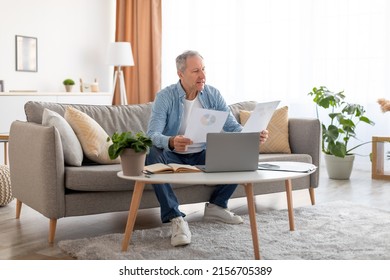 Mature Male Self-Entrepreneur Working With Papers At Desk In Home Office Sitting On Sofa, Checking Annual Financial Reports. Busy Man Reading Documents, Sitting At Table With Pc Computer, Copy Space