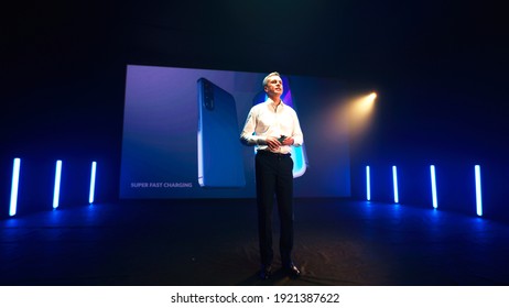 Mature Male Speaker In Smart Casual Clothes Walking On Stage And Showing New Smartphone Against LED Screen