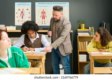 Mature Male Teacher Of Anatomy Checking Notes Of Highschool Student And Consulting Him Among Classmates At Lesson In Classroom