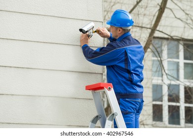 Mature Male Technician Standing On Stepladder Fitting CCTV Camera On Wall