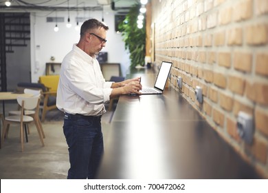 Mature Man 40 Years Old In Eyeglasses Working At Modern Netbook Computer Connecting To Wireless 4G Internet In Coworking Space.Entrepreneur Standing At Digital Laptop And Checking Online Database