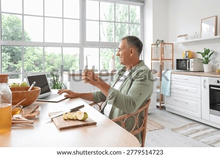 Mature man with apple cider using laptop at table in kitchen