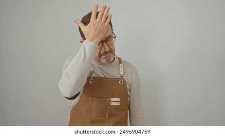 A mature man in an apron looking frustrated, touches his head against a white background. - Powered by Shutterstock