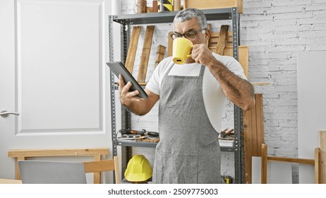 A mature man in an apron sips coffee while browsing a tablet in a well-equipped workshop. - Powered by Shutterstock