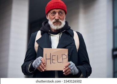 Mature Man Asking For Some Help Standing On City Street. Homeless Man In Warm Coat And Red Hat