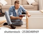 Mature Man Assembling Shelf Using Electric Drill Tool Sitting On Floor At Home. Male Housework And House Furnishing. Handyman Fixing Wooden Cabinet During Renovation