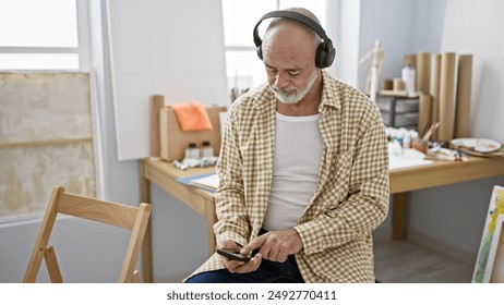 A mature man with beard and headphones using a smartphone indoors at a studio with art supplies. - Powered by Shutterstock