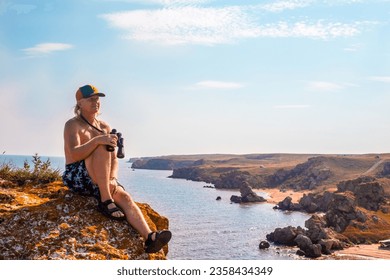 Mature man with binoculars sits on a rock on top of a mountain and looks at the seascape. Rocky coast of the Sea of Azov, General's beaches. Travel and tourism. - Powered by Shutterstock