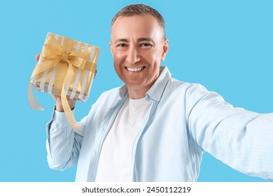 Mature man with birthday gift taking selfie on blue background, closeup - Powered by Shutterstock