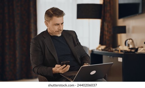 Mature man in a business jacket multitasking with a laptop computer and smartphone in a hotel room. Businessman working on business trip. - Powered by Shutterstock