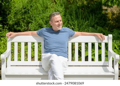 A mature man in casual attire sits on a white bench, reflecting and enjoying the peaceful surroundings of a lush park. - Powered by Shutterstock