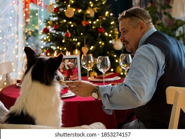 Mature man celebrating Christmas with his dog sitting at served holiday  table with laptop. People greeting their friends on video call using webcam. Christmas eve online. New normal social distancing