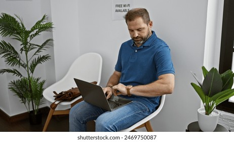 Mature man checking watch while working on laptop in a modern indoor lobby with plants and free wi-fi sign in background.
