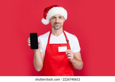 Mature Man In Christmas Santa Hat And Red Apron Hold Smartphone And Card. Online Banking