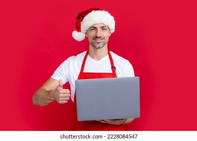 mature man in christmas santa hat and red apron hold laptop. thumb up - Powered by Shutterstock