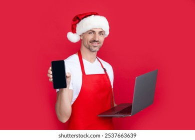 mature man in christmas santa hat and red apron hold laptop with phone - Powered by Shutterstock