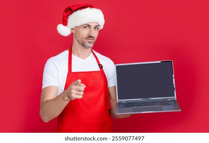 mature man in christmas santa hat and red apron presenting laptop. point finger - Powered by Shutterstock