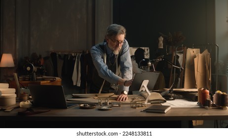 Mature man creates clothes design for new wear collection using tablet and laptop. Tailor in glasses looks at camera, stands near table with tailoring tools. Concept of fashion and hand craft. - Powered by Shutterstock