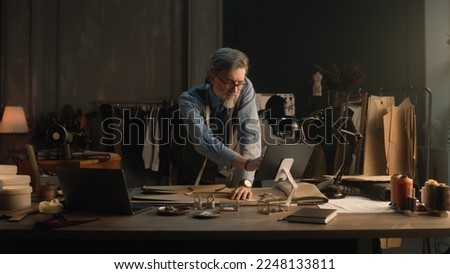 Mature man creates clothes design for new wear collection using tablet and laptop. Tailor in glasses looks at camera, stands near table with tailoring tools. Concept of fashion and hand craft.