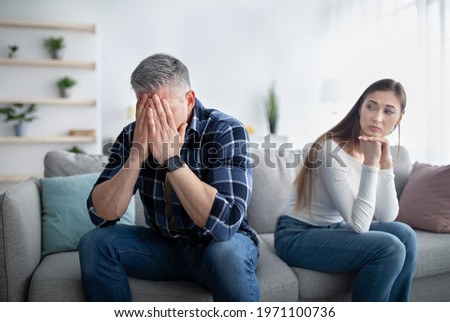 Mature man crying on sofa, his upset wife looking at him, indoors. Middle-aged couple suffering from difficulties in relationship, sitting separated on couch in living room