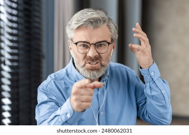 Mature man displaying anger and frustration during virtual meeting in modern office setting. Wearing glasses, gray beard, blue shirt, expressing emotions. Intense expression and gestures. - Powered by Shutterstock