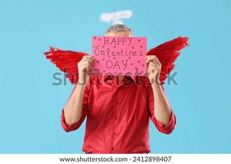 Mature man dressed as Cupid with card on blue background. Valentine's Day celebration