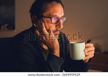Mature man drinking coffee while looking at computer screen in dark room with warm light and low key atmosphere. Selective focus.