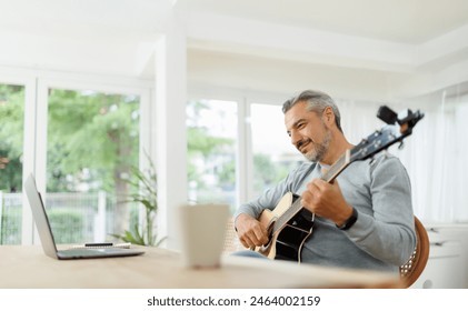 Mature man enjoying a guitar session in a white room with a laptop and coffee mug on the table. - Powered by Shutterstock