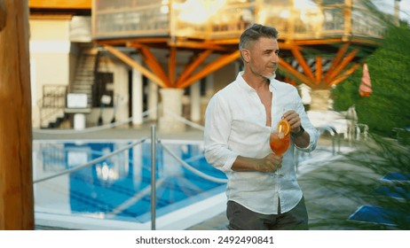 Mature man enjoying summer vacation in holiday resort. Handsome older male holding refreshing aperitivo cocktail by poolside, smiling with satisfaction. - Powered by Shutterstock