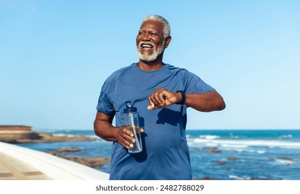 Mature man exercising at the seaside, wearing a fitness watch. He is holding a water bottle and looking enthusiastic and healthy - focusing on longevity. - Powered by Shutterstock